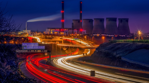 Nighttime long-exposure photograph of a multi-lane highway curving toward cooling towers and tall smokestacks.